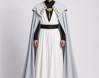 Puede incluir: Imagen de cuerpo entero de una persona con un atuendo de inspiración fantástica. El conjunto incluye una capa gris claro con botones dorados, un vestido blanco, una parte superior negra y un cinturón negro. Un colgante dorado cuelga del escote. La persona lleva botas marrones.