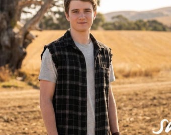 Puede incluir: Una persona viste una camiseta gris debajo de una camisa sin mangas a cuadros negros y grises con sisas deshilachadas. La palabra SWAG está impresa en blanco en la esquina inferior derecha de la imagen sobre un fondo de campo rural.