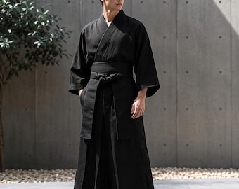 Puede incluir: Una túnica negra de estilo kimono con mangas anchas y cierre cruzado en la parte delantera, asegurada con una faja de tela ancha a juego atada en un nudo en la cintura. La prenda incluye pantalones largos y fluidos con aspecto plisado.