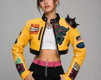 Puede incluir: Chaqueta corta de cuero amarilla con detalles negros, parches y una hombrera decorativa. La chaqueta tiene el texto "Rrrrrrumi" y "HUNTR". La modelo lleva pantalones cargo negros y un cinturón rosa.