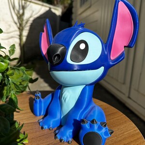 Puede incluir: Figura azul del personaje Stitch de la pel&iacute;cula de animaci&oacute;n Lilo & Stitch. La figura tiene orejas grandes con interiores rosas, una nariz negra y ojos grandes. Est&aacute; sobre una superficie de madera.