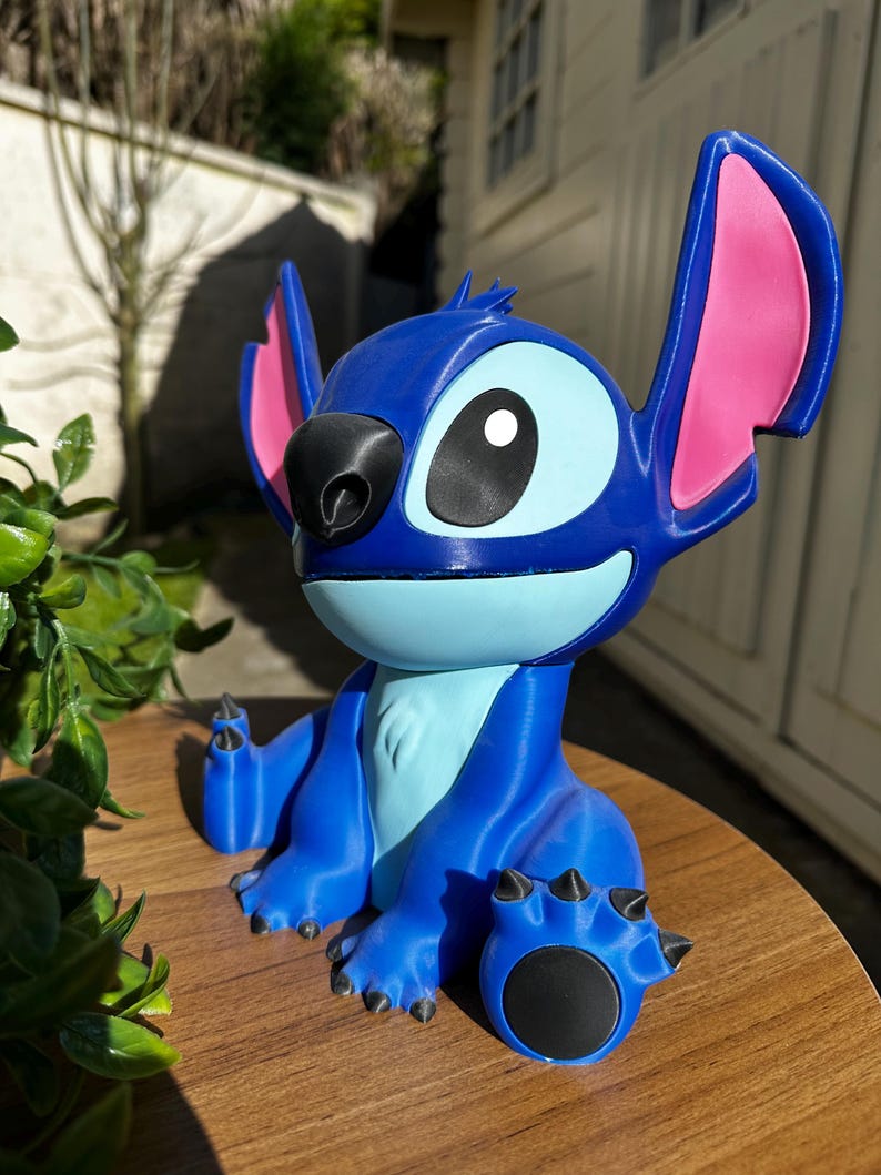 Puede incluir: Figura azul del personaje Stitch de la pel&iacute;cula de animaci&oacute;n Lilo & Stitch. La figura tiene orejas grandes con interiores rosas, una nariz negra y ojos grandes. Est&aacute; sobre una superficie de madera.