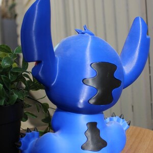 Puede incluir: Figura azul de Stitch, con grandes orejas, manchas negras y una espalda puntiaguda. La figura est&aacute; sentada, con una boca peque&ntilde;a y un dise&ntilde;o detallado. La figura es de pl&aacute;stico.