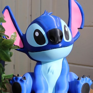 Puede incluir: Figura azul del personaje Stitch de la pel&iacute;cula de animaci&oacute;n Lilo & Stitch. La figura tiene orejas grandes con interior rosa, una nariz negra y ojos grandes y negros. La figura est&aacute; sentada con los brazos y las piernas extendidos.