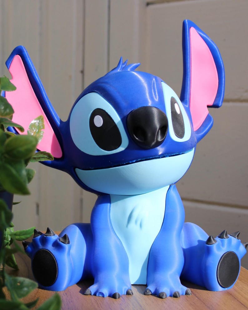 Puede incluir: Figura azul del personaje Stitch de la pel&iacute;cula de animaci&oacute;n Lilo & Stitch. La figura tiene orejas grandes con interior rosa, una nariz negra y ojos grandes y negros. La figura est&aacute; sentada con los brazos y las piernas extendidos.