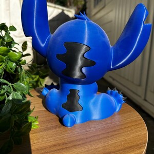 Puede incluir: Figura azul de un personaje de dibujos animados con grandes orejas y una mancha negra en el pecho. La figura est&aacute; sobre una superficie de madera. El personaje tiene una cabeza redonda y detalles puntiagudos.