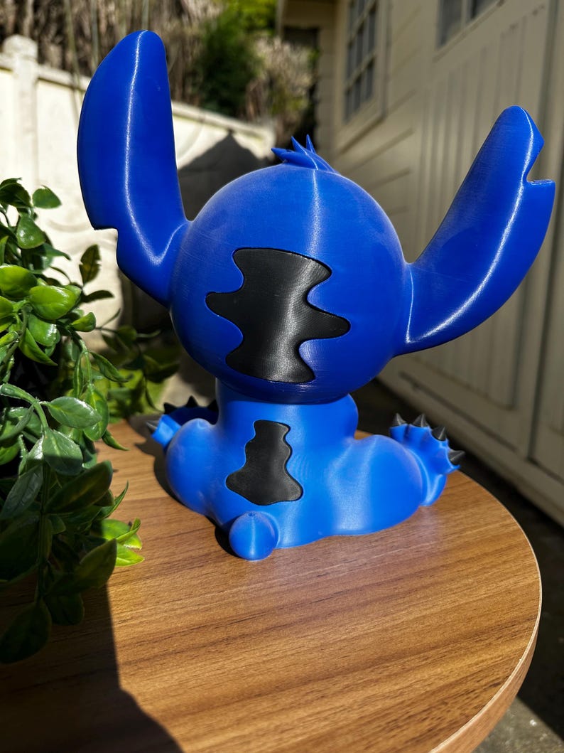 Puede incluir: Figura azul de un personaje de dibujos animados con grandes orejas y una mancha negra en el pecho. La figura est&aacute; sobre una superficie de madera. El personaje tiene una cabeza redonda y detalles puntiagudos.