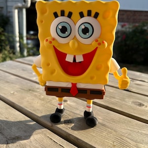 Pode incluir: Uma figura retangular amarela do Bob Esponja com olhos azuis grandes, uma boca larga sorridente e dois dentes da frente. O personagem veste uma camisa branca, uma gravata vermelha, calças castanhas e sapatos pretos, fazendo um gesto de positivo.
