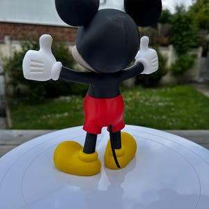 K&ouml;nnte beinhalten: Eine 3D-gedruckte Mickey Mouse-Figur, von hinten gesehen, mit erhobenen Armen und Daumen hoch. Die Figur hat einen schwarzen Kopf mit gro&szlig;en Ohren, wei&szlig;e Handschuhe, rote Shorts und gelbe Schuhe. Auf einer wei&szlig;en Oberfl&auml;che.