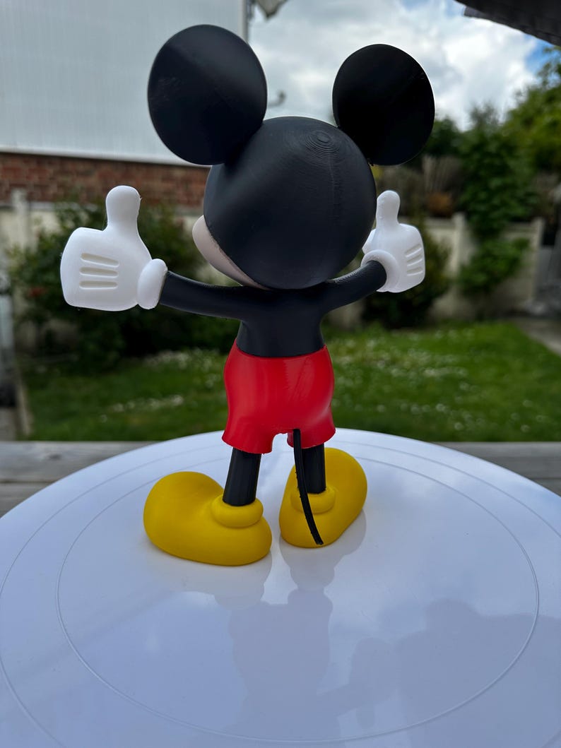 K&ouml;nnte beinhalten: Eine 3D-gedruckte Mickey Mouse-Figur, von hinten gesehen, mit erhobenen Armen und Daumen hoch. Die Figur hat einen schwarzen Kopf mit gro&szlig;en Ohren, wei&szlig;e Handschuhe, rote Shorts und gelbe Schuhe. Auf einer wei&szlig;en Oberfl&auml;che.