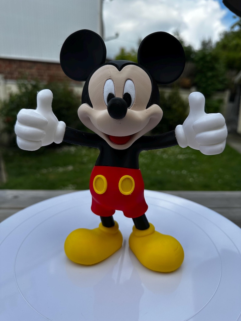 K&ouml;nnte beinhalten: Eine Mickey Mouse-Figur, eine klassische Zeichentrickfigur, mit schwarzen Ohren, einem schwarzen K&ouml;rper und wei&szlig;en Handschuhen. Er tr&auml;gt rote Shorts mit gelben Kn&ouml;pfen und gelbe Schuhe und zeigt den Daumen nach oben.
