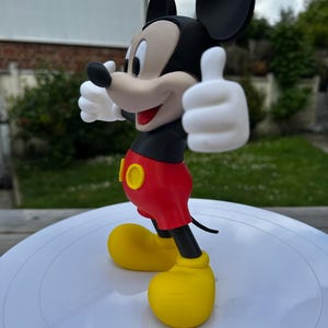 K&ouml;nnte beinhalten: Eine Mickey Mouse-Figur mit erhobenem Daumen. Die Figur hat einen schwarzen Kopf mit gro&szlig;en Ohren, ein beiges Gesicht und wei&szlig;e Handschuhe. Sie tr&auml;gt ein schwarzes Oberteil, rote Shorts und gelbe Schuhe. Die Figur steht auf einer wei&szlig;en Oberfl&auml;che.