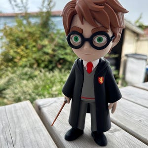 Peut inclure: Une figurine stylisée d'une jeune personne aux cheveux bruns, portant des lunettes rondes noires, un pull gris, une cravate rouge et une robe noire avec un écusson jaune et rouge. La figurine tient une baguette brune fine et a une cicatrice.