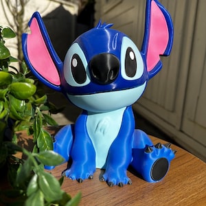 Puede incluir: Una figura azul de un personaje de dibujos animados con orejas grandes, una nariz negra y ojos grandes. La figura tiene detalles rosas en las orejas y un vientre azul claro. Est&aacute; sobre una superficie de madera con follaje verde en primer plano.