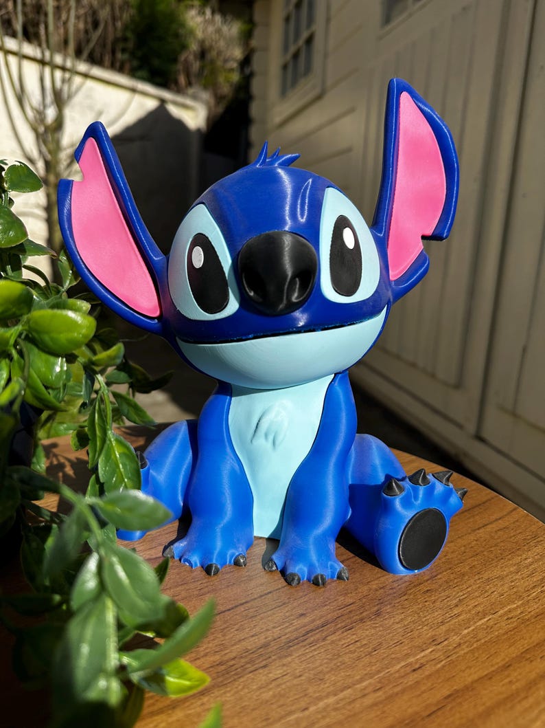Puede incluir: Una figura azul de un personaje de dibujos animados con orejas grandes, una nariz negra y ojos grandes. La figura tiene detalles rosas en las orejas y un vientre azul claro. Est&aacute; sobre una superficie de madera con follaje verde en primer plano.