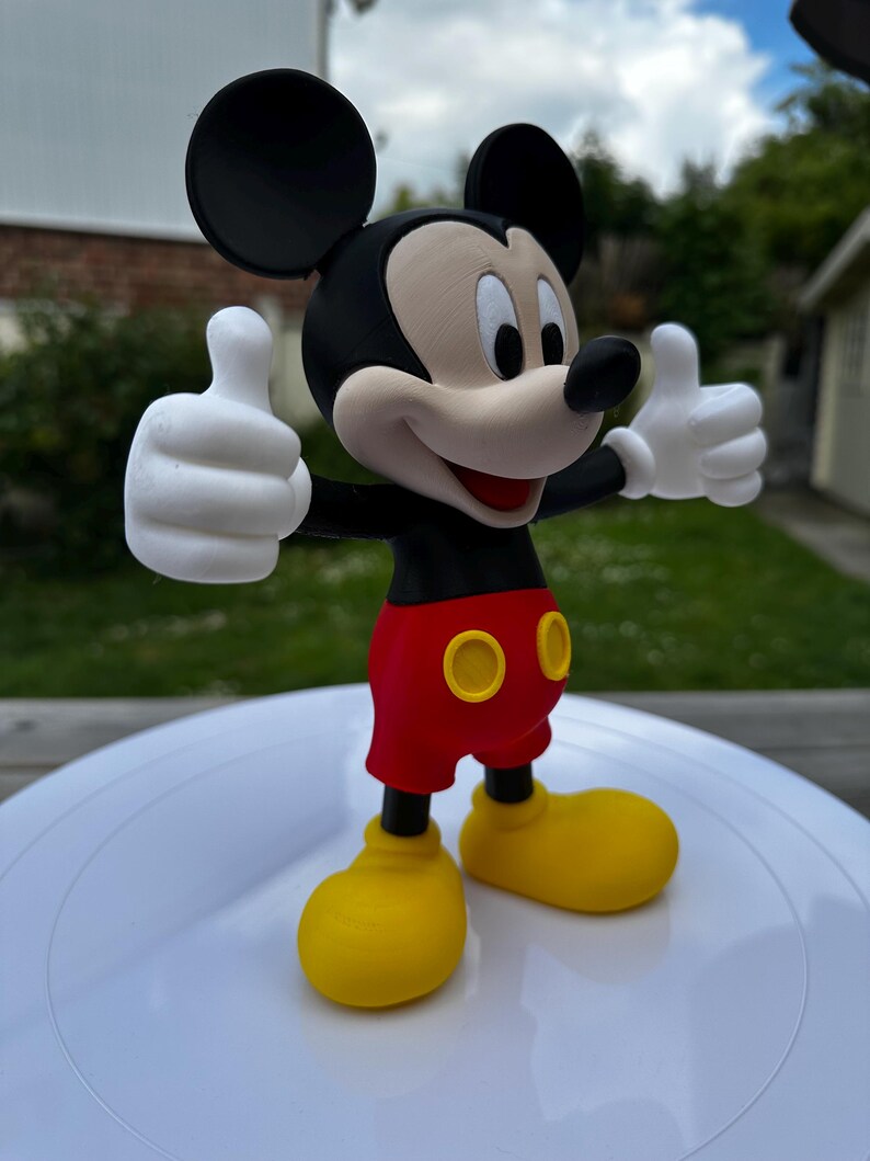 K&ouml;nnte beinhalten: Eine 3D-gedruckte Mickey Mouse-Figur. Die Figur hat schwarze Ohren, einen schwarzen Torso, rote Shorts mit gelben Kn&ouml;pfen, gelbe Schuhe und wei&szlig;e Handschuhe. Mickey hebt beide Daumen.
