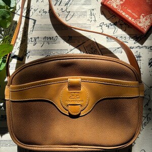 May include: A tan, textured leather crossbody bag with a rounded silhouette and a long shoulder strap. The front features a smooth leather horizontal band with a central circular tab embossed with the Courr&egrave;ges logo and the text courr&egrave;ges.