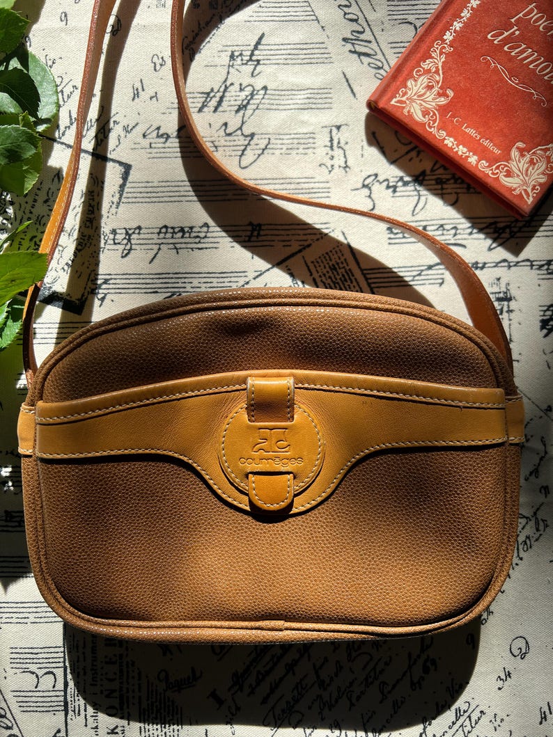 May include: A tan, textured leather crossbody bag with a rounded silhouette and a long shoulder strap. The front features a smooth leather horizontal band with a central circular tab embossed with the Courr&egrave;ges logo and the text courr&egrave;ges.