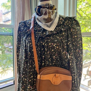 May include: A dark green long-sleeved dress with a small floral pattern, featuring a ruffled yoke and collar. A brown leather crossbody bag with a tan leather trim and a central clasp is worn over the shoulder. A white silk scarf with a black and gold tassel print is tied around the neck of the mannequin.
