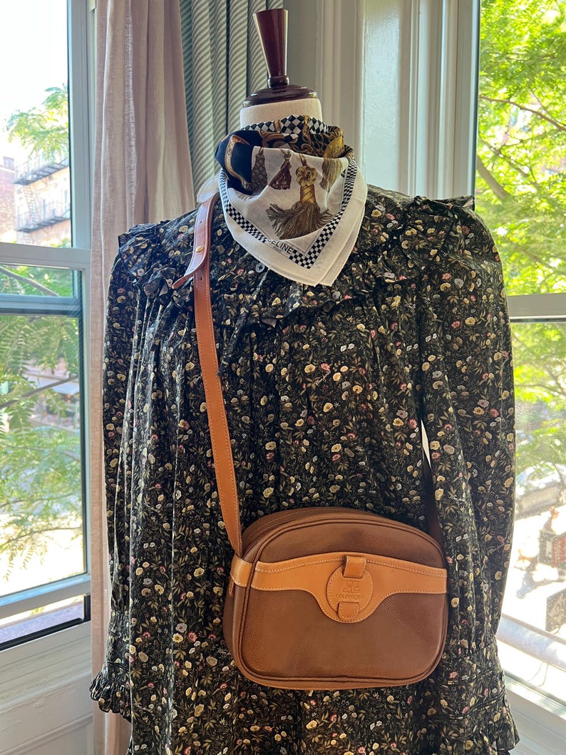May include: A dark green long-sleeved dress with a small floral pattern, featuring a ruffled yoke and collar. A brown leather crossbody bag with a tan leather trim and a central clasp is worn over the shoulder. A white silk scarf with a black and gold tassel print is tied around the neck of the mannequin.
