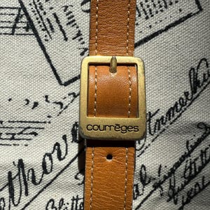 May include: A tan leather strap featuring a rectangular gold-toned metal buckle with the word courr&egrave;ges engraved on the front. The strap has visible white stitching along the edges and a single adjustment hole visible below the buckle.