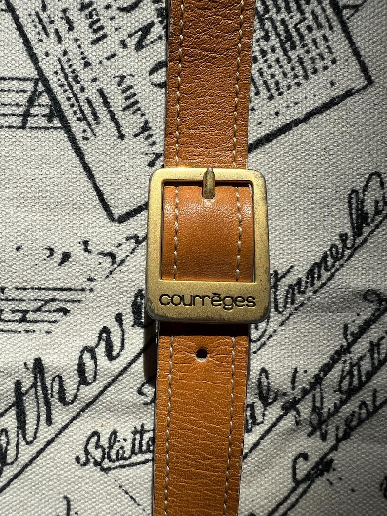 May include: A tan leather strap featuring a rectangular gold-toned metal buckle with the word courr&egrave;ges engraved on the front. The strap has visible white stitching along the edges and a single adjustment hole visible below the buckle.