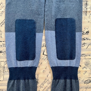 Puede incluir: Primer plano de un pantalón de chándal azul y gris. El pantalón presenta un diseño de bloques de color con parches en las rodillas de color azul oscuro y puños acanalados. La tela parece ser un tejido de punto suave.