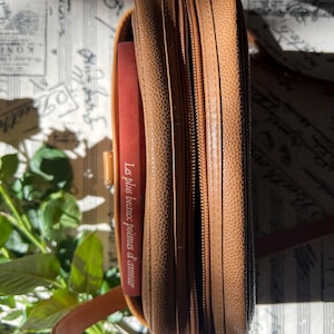 May include: A tan textured leather crossbody bag with a zippered closure and a thin shoulder strap. Inside the bag, a red book spine is visible with the white text Les plus beaux po&egrave;mes d'amour printed vertically along the side of the cover.