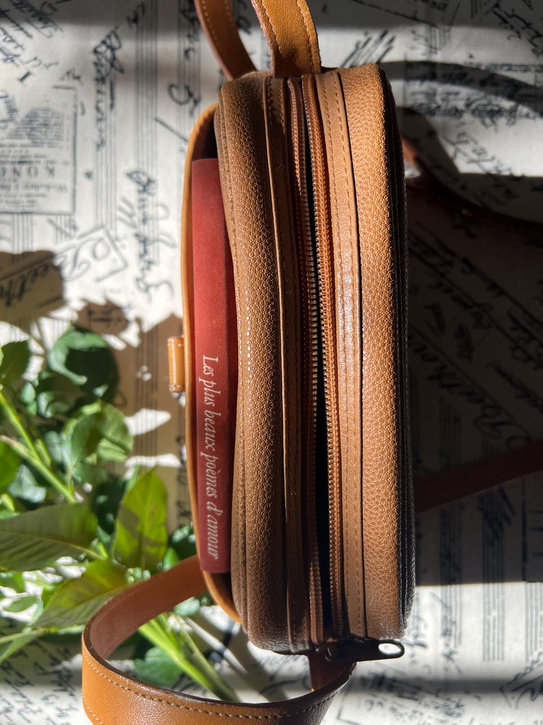 May include: A tan textured leather crossbody bag with a zippered closure and a thin shoulder strap. Inside the bag, a red book spine is visible with the white text Les plus beaux po&egrave;mes d'amour printed vertically along the side of the cover.