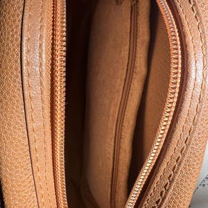 May include: A tan-colored bag with a textured exterior and a matching tan interior lining. The bag features a top zipper closure with a metal pull and an internal zippered pocket located on the side wall, providing a secure compartment for small items.