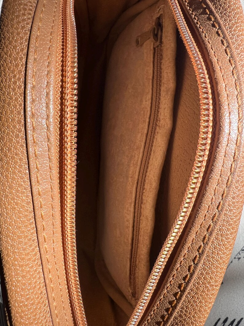 May include: A tan-colored bag with a textured exterior and a matching tan interior lining. The bag features a top zipper closure with a metal pull and an internal zippered pocket located on the side wall, providing a secure compartment for small items.