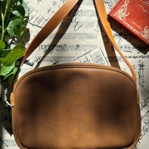 May include: A medium-sized, oval-shaped shoulder bag in a solid tan color with a textured, pebbled surface. The bag features a matching tan shoulder strap attached to the sides and a zipper closure along the top edge, maintaining a clean, simple design.