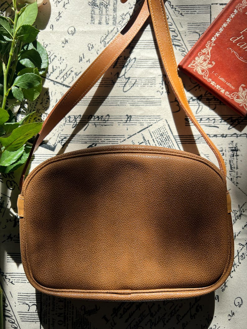 May include: A medium-sized, oval-shaped shoulder bag in a solid tan color with a textured, pebbled surface. The bag features a matching tan shoulder strap attached to the sides and a zipper closure along the top edge, maintaining a clean, simple design.