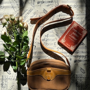 May include: A tan leather crossbody bag with a rounded silhouette, a front flap pocket, and an adjustable shoulder strap. Beside the bag is a bouquet of pale pink roses and a red book titled Les plus beaux po&egrave;mes d'amour with gold lettering.