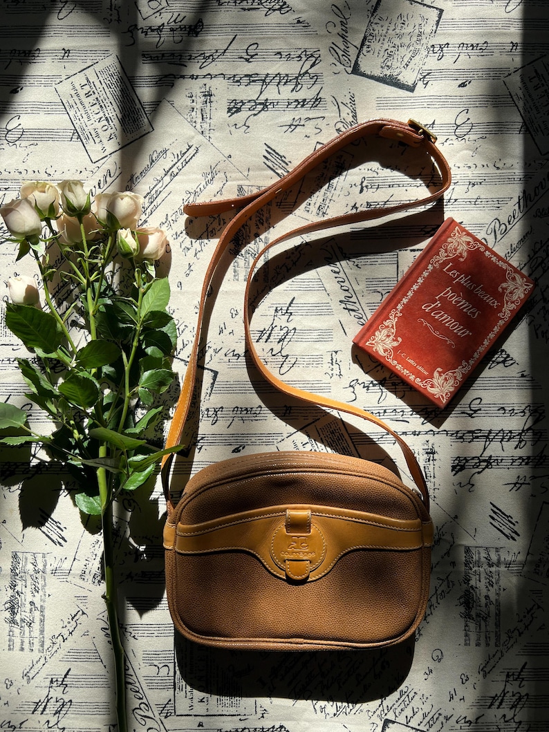 May include: A tan leather crossbody bag with a rounded silhouette, a front flap pocket, and an adjustable shoulder strap. Beside the bag is a bouquet of pale pink roses and a red book titled Les plus beaux po&egrave;mes d'amour with gold lettering.
