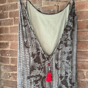 May include: A sleeveless dress featuring a grey and dark brown snakeskin print pattern. The garment has a deep V-shaped back neckline with thin spaghetti straps and a bright red tassel accent hanging from the centre of the back strap detail.