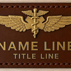 Peut inclure: Une étiquette de nom en cuir marron avec des lettres dorées et un emblème doré représentant des ailes, un caducée et une croix. Le texte sur l'étiquette indique "NAME LINE" et "TITLE LINE". Les bords sont cousus.
