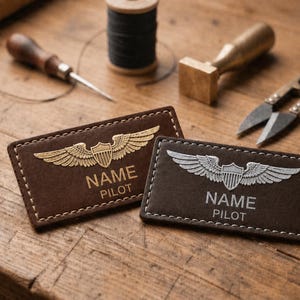 May include: Two brown leather pilot name tags with stitched edges, featuring a wing and shield emblem. One tag has gold-colored details, the other silver. The text "NAME" and "PILOT" are visible. Crafting tools are in the background.