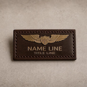 Peut inclure: Un porte-nom rectangulaire en cuir marron avec des bords cousus. Il présente un emblème doré d'ailes et d'un bouclier. Les textes "NAME LINE" et "TITLE LINE" sont gravés en lettres dorées.
