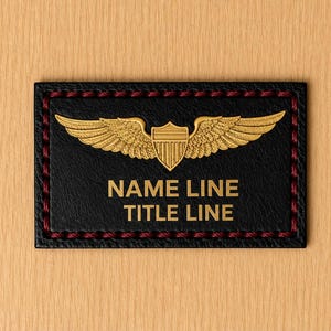 May include: A rectangular black leather patch featuring a gold-coloured metallic emblem of wings flanking a shield. Below the emblem, the text reads NAME LINE and TITLE LINE in gold. The patch is bordered by dark red stitching along the edges.