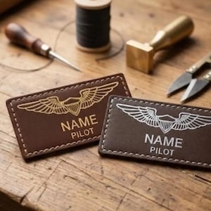 May include: Two brown leather pilot name tags with embroidered wings and the text "NAME PILOT". One tag has gold embroidery, the other silver. Crafting tools, including a needle, thread, and scissors, are in the background.