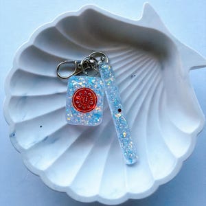 Könnte beinhalten: Ein Schlüsselanhänger mit zwei Harzanhängern, die mit irisierendem blauem und weißem Glitzer gefüllt sind. Ein Anhänger ist ein rechteckiger Mahjong-Stein mit einem roten kreisförmigen Symbol, der andere ein langer, dünner, abgerundeter Stab. Beide an einem silbernen Metallverschluss.