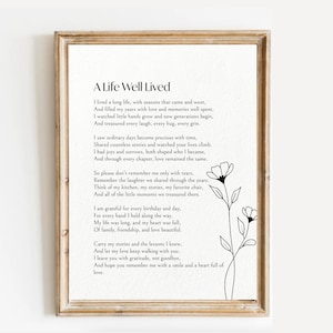 May include: A framed print featuring the title A Life Well Lived in black text on a white background. The poem describes a long life filled with memories, love, and family. A simple black line drawing of a flower stem with two blossoms is on the right.