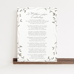 Op de afbeelding: Een ingelijste print met het gedicht "A Mother's Love, Everlasting", omgeven door een bloemenrand. De tekst staat in sierlijke letters op een crèmekleurige achtergrond, gepresenteerd op een houten plank.