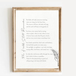 May include: A framed print with a light beige background features a poem titled "The Fields Still Remember" in elegant black script. The poem is about farming and nature, with a wheat illustration in the bottom right corner. The frame is a light brown colour.