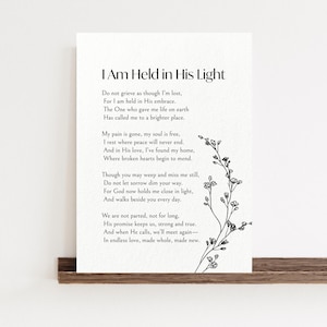 May include: A white print featuring the text "I Am Held in His Light" and a poem. A delicate black floral illustration is on the right side. The print is displayed on a wooden shelf.