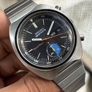May include: A vintage Seiko 5 Sports SpeedTimer watch with a black dial, silver case, and a stainless steel strap. The watch features a chronograph function with an orange second hand and a blue subdial. The day and date are displayed at the 3 o'clock position.
