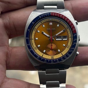 May include: A vintage Seiko chronograph automatic watch with a gold-toned dial, blue and red accents, and a stainless steel band. The watch face displays the day and date. The watch has a tachymeter scale.