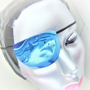 Op de afbeelding: Een blauw en wit ooglapje met een haai-ontwerp. Het ooglapje is gemaakt van stof en heeft een elastisch bandje.