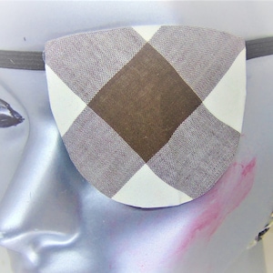 Puede incluir: Un parche para el ojo de cuadros marrones y blancos con una banda elástica negra. El parche para el ojo se lleva en una cabeza de maniquí.